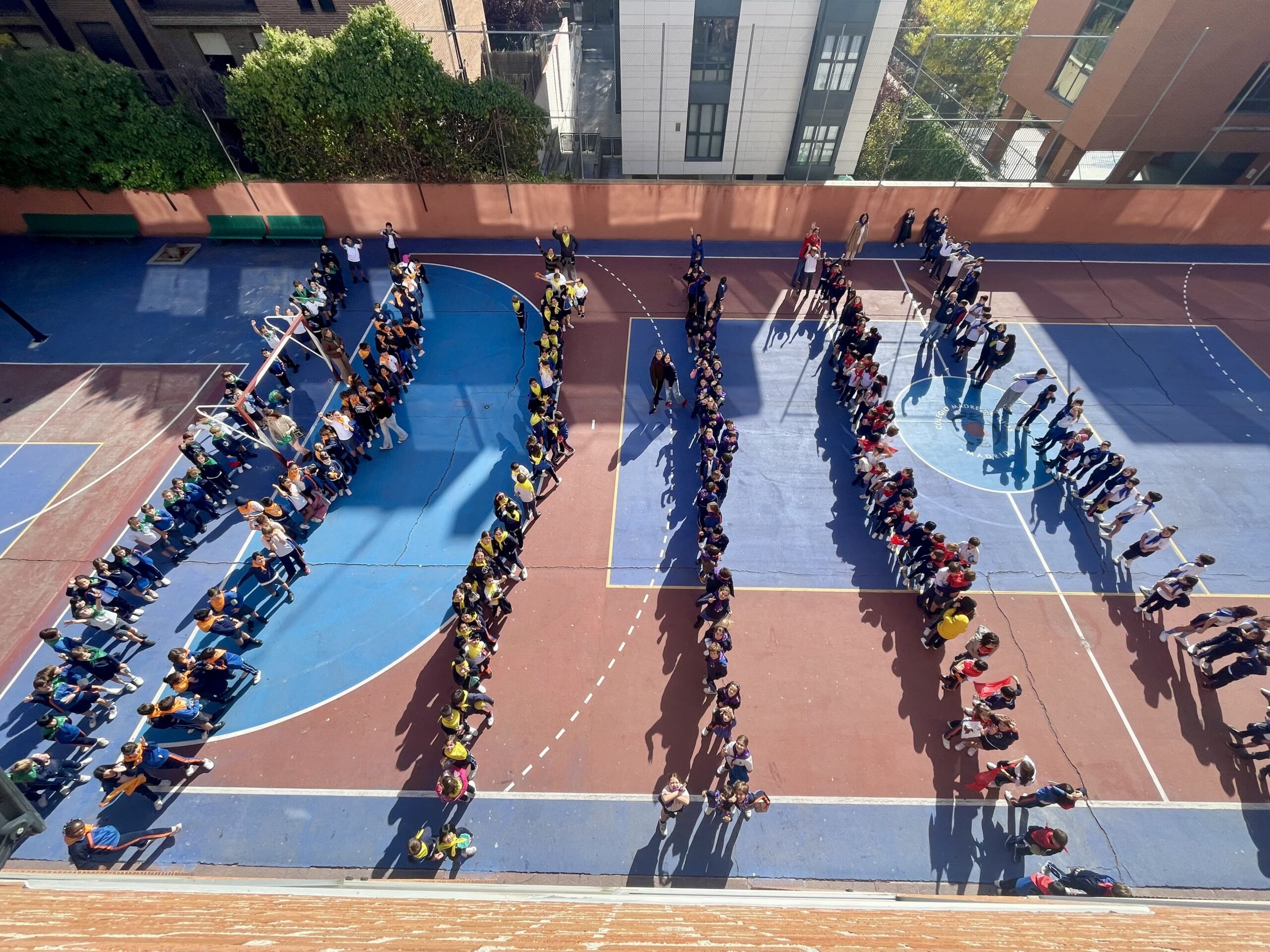 Vista aérea de estudiantes formando filas organizadas en una cancha de baloncesto de una escuela, formando la palabra FELIZ con sus cuerpos. La cancha está dividida en secciones azules y rojas, rodeadas de edificios y vegetación.