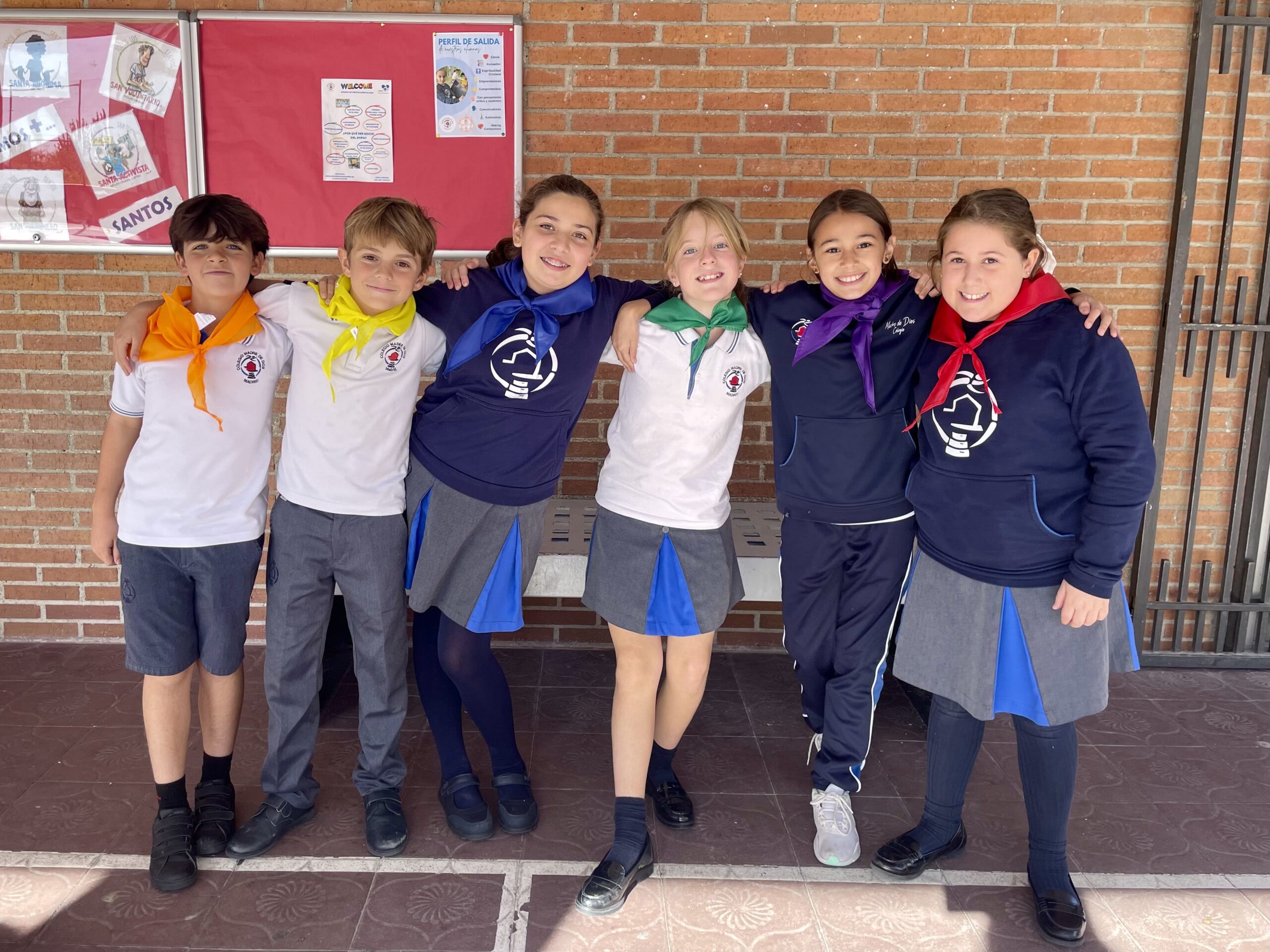 Seis niños sonrientes con uniforme escolar permanecen de pie, cogidos del brazo, frente a una pared de ladrillo y un tablón de anuncios. Cada niño lleva un pañuelo de distinto color alrededor del cuello.