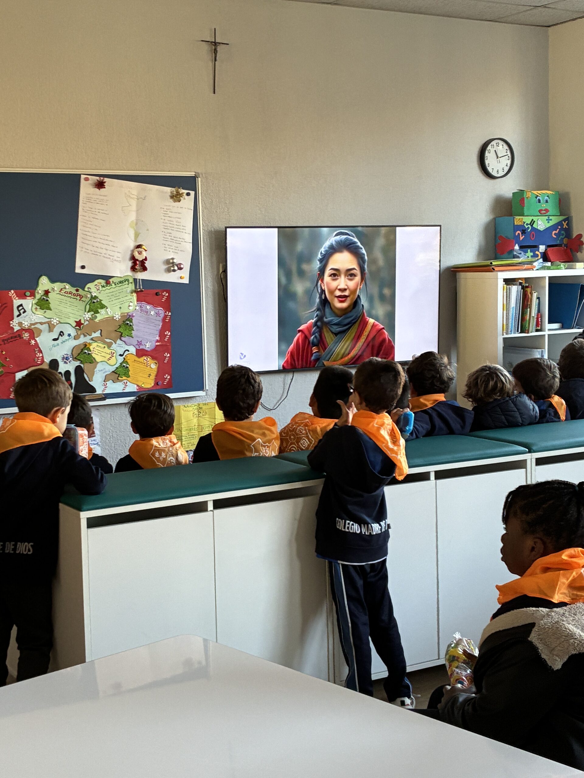 Un grupo de niños pequeños en un aula observan a una mujer que habla en una pantalla de televisión. El aula tiene carteles educativos, un reloj y coloridos adornos en las paredes.