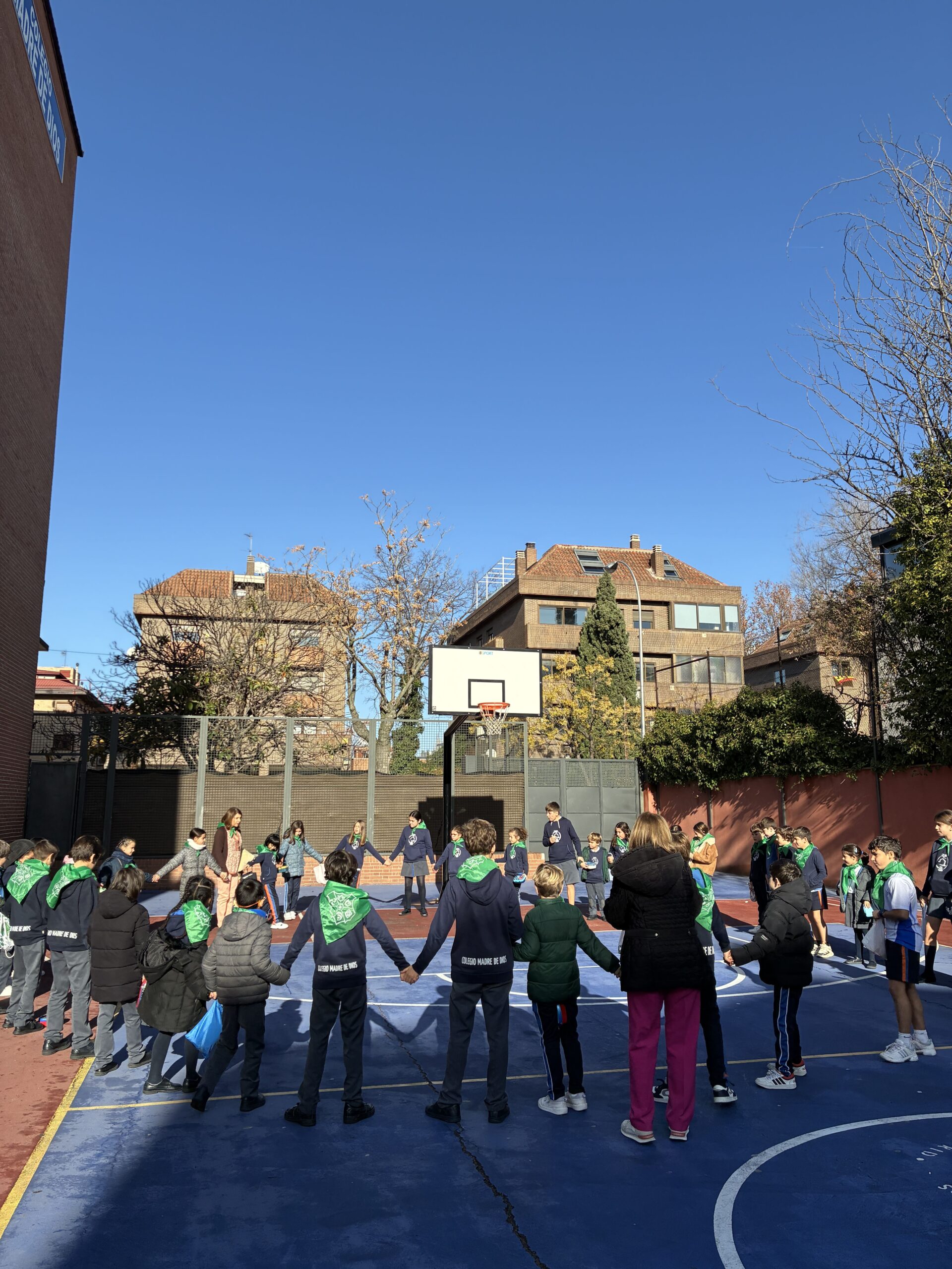 Un grupo de niños y algunos adultos forman un gran círculo en una cancha de baloncesto al aire libre, sujetando un gran paracaídas. El cielo está despejado y azul, y al fondo se ven edificios y árboles.